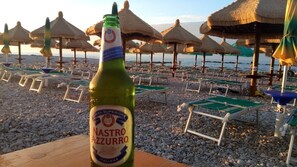 A bottle of Nastro Azzurro on a table with beach chairs and umbrellas in the background.