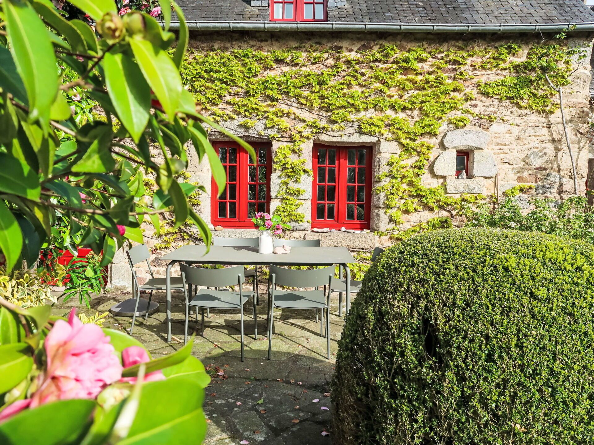 Un bâtiment en pierre avec des portes et des fenêtres rouges, un mur de pierre recouvert de lierre et une table avec des chaises devant.