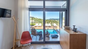 Una habitación de hotel moderna con un amplio ventanal corredero que da a una zona de piscina, un escritorio de madera, una silla roja y un televisor montado en la pared.