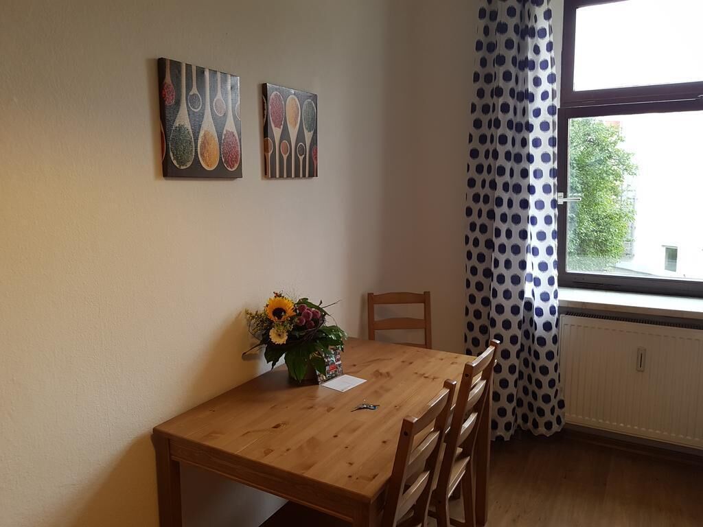 Ein Essbereich mit einem Holztisch, Stühlen und einer Vase mit Blumen. Zwei Gemälde hängen an der Wand, und ein Fenster ist mit Vorhängen drapiert.