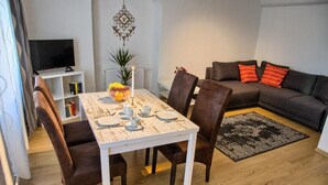 Een eetgedeelte met een witte tafel voor vier personen, omringd door donkerbruine stoelen. Links is een televisie bevestigd aan een wit schap, en op de achtergrond staat een grijze bank met rode kussens.