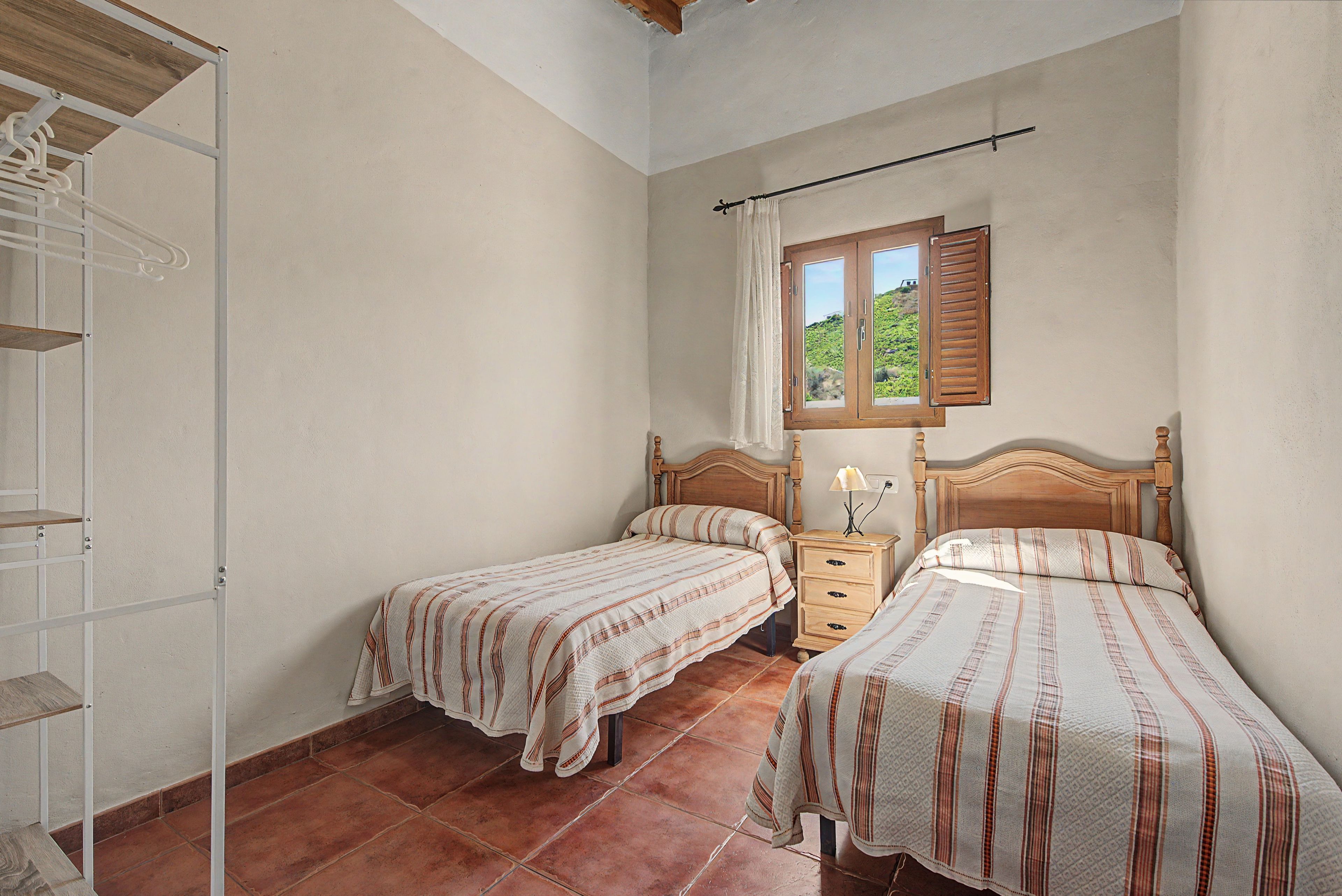 A bedroom with two single beds, a nightstand, a window with shutters, and a wooden ceiling.