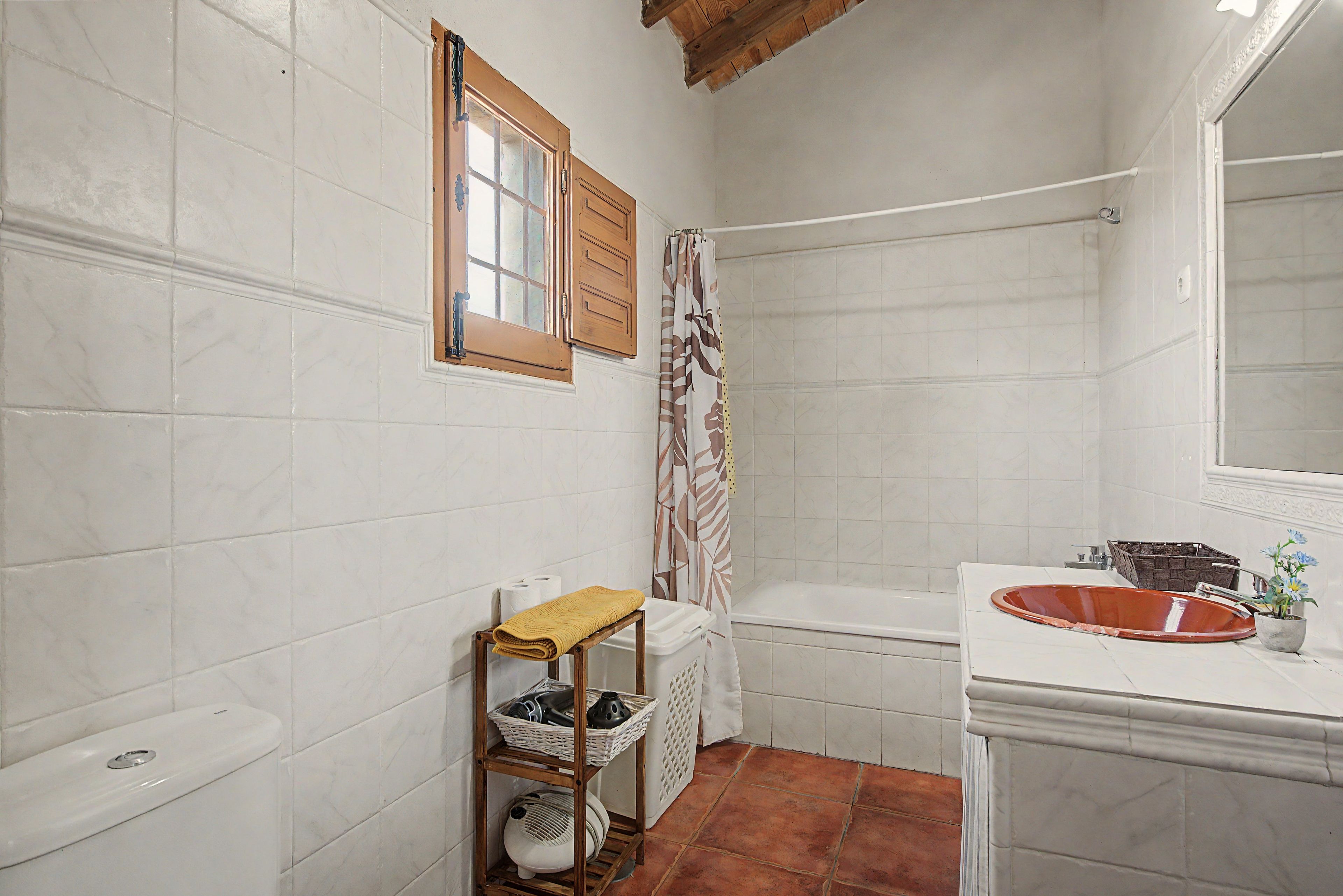A bathroom with white tiled walls, a wooden shelf with baskets, a white bathtub, and a wooden window with shutters.