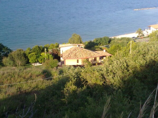 A coastal house with a tiled roof, surrounded by greenery and overlooking the sea.