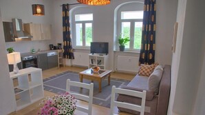 A modern living room with a sofa, coffee table, and TV. There is a kitchen area with cabinets and a stove. Large windows with curtains are visible. - Dresdner-Tag Helene (Dresden)