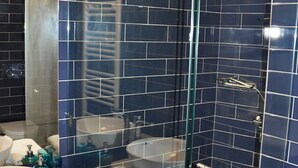 Un baño con azulejos azul oscuro, ducha con cerramiento de vidrio, lavamanos blanco y un espejo.