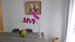 Una mesa de comedor con un jarrón de flores moradas, un frutero y un cuadro en la pared.