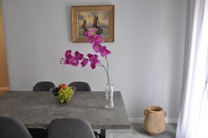 Une table à manger ornée d’un vase de fleurs violettes, d’un saladier de fruits et d’un tableau accroché au mur.
