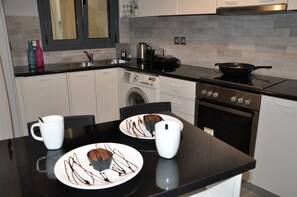 Une cuisine moderne avec un plan de travail noir, des placards blancs, un lave-linge, un four et un coin repas avec des assiettes remplies de nourriture et des tasses.
