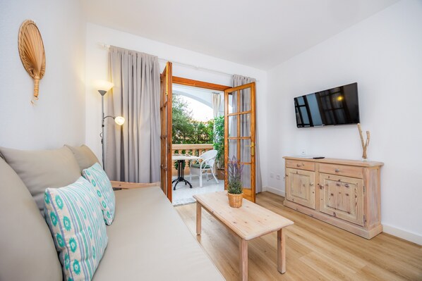 Een moderne woonkamer met een bank, een houten tafel, een aan de muur bevestigde televisie en uitzicht door een open deur naar een buitenruimte.
