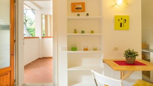 A room with a wooden table, white chairs, a red placemat, a potted plant, and shelves with various items. - Studio near the beach - Apartment Lucia (Campo nell'Elba)