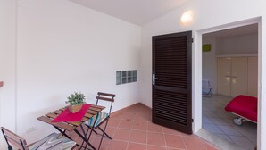 A room with a tiled floor, a wooden table with a red cloth, a chair, a small plant, a door, and a bed with a red cover. - Studio near the beach - Apartment Lucia (Campo nell'Elba)