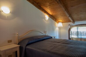 Un dormitorio con techo de madera, una cama con cubrecama acolchado, una mesita de noche, una lámpara y una ventana con cortinas.