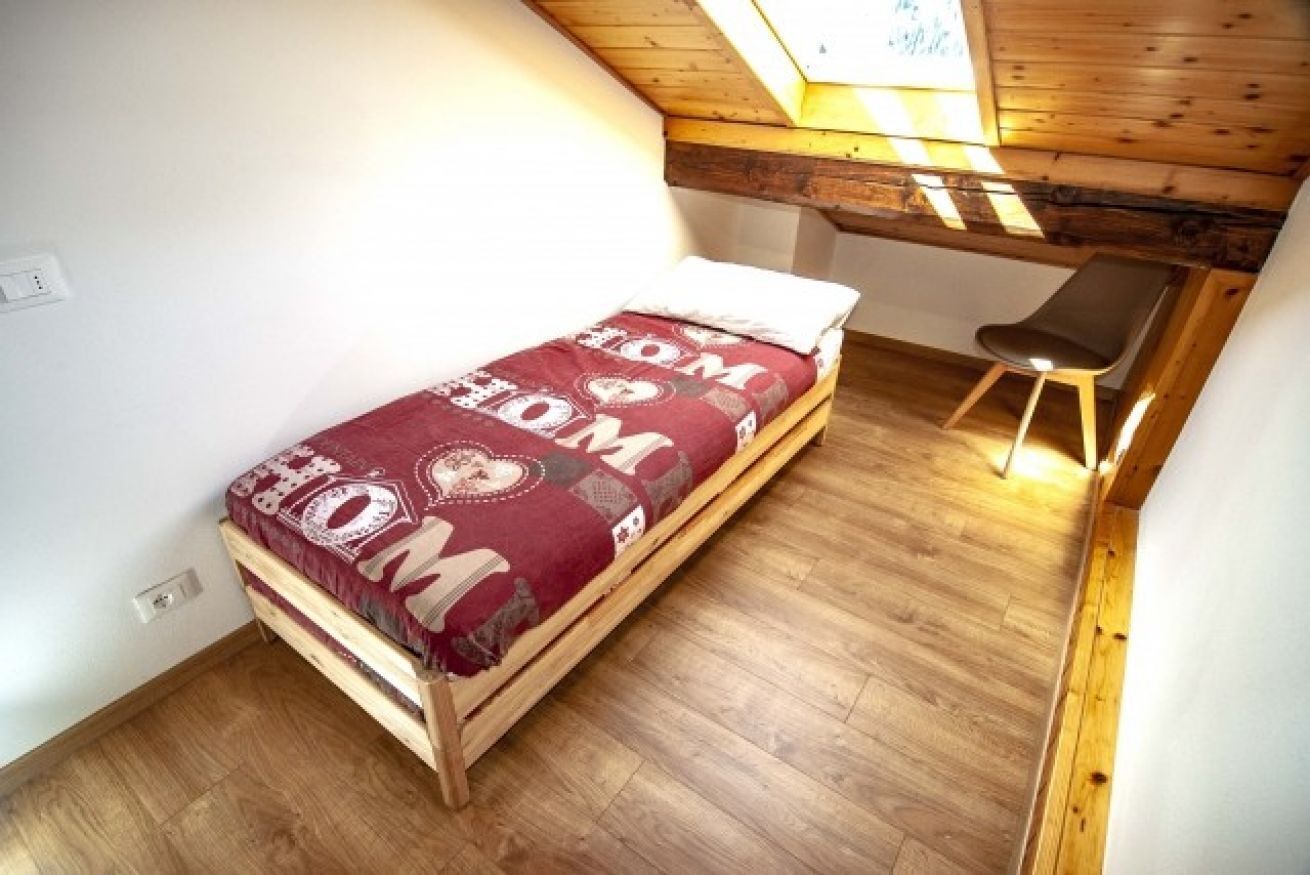 A small, single-bed room with a sloped ceiling and wooden beams, a bench, and a skylight.