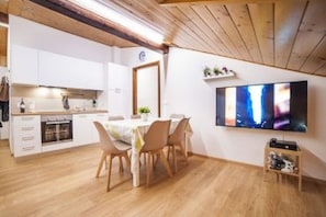A modern kitchen with white cabinets, a dining area with a table and chairs, a TV mounted on the wall, and a wooden ceiling.