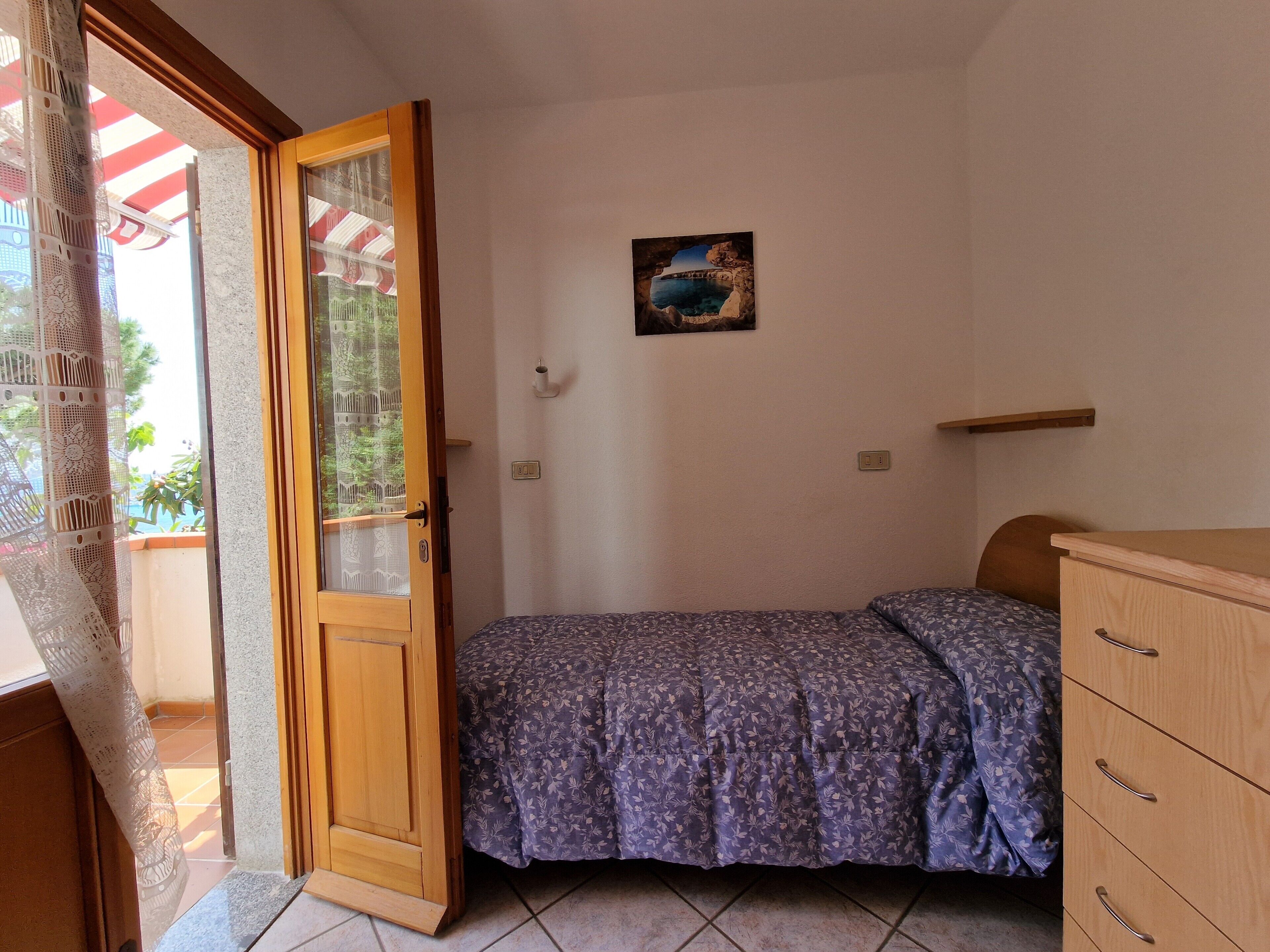 Ein Schlafzimmer mit einem Bett, einer Holzkommode, einem Bild an der Wand und einer Tür zu einem Außenbereich.