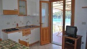 Een kleine keuken met een spoelbak, fornuis en houten kastjes, een eethoek met een tafel en stoelen, en een balkon met een televisie.