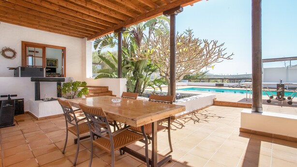Een overdekt terras met een houten tafel en stoelen, een zwembad op de achtergrond en uitzicht op het omliggende landschap.