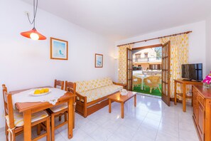 A living room with a sofa, dining table, chairs, a TV, and a balcony with a view. - Apartment Close to the Beach with Pool Access, Private Terrace, Air Conditioning and Wi-Fi (Canyamel)