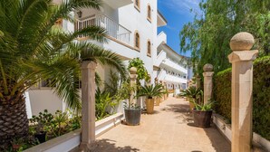 A corridor with potted plants, a palm tree, and white buildings with balconies. - Apartment Close to the Beach with Pool Access, Private Terrace, Air Conditioning and Wi-Fi (Canyamel)