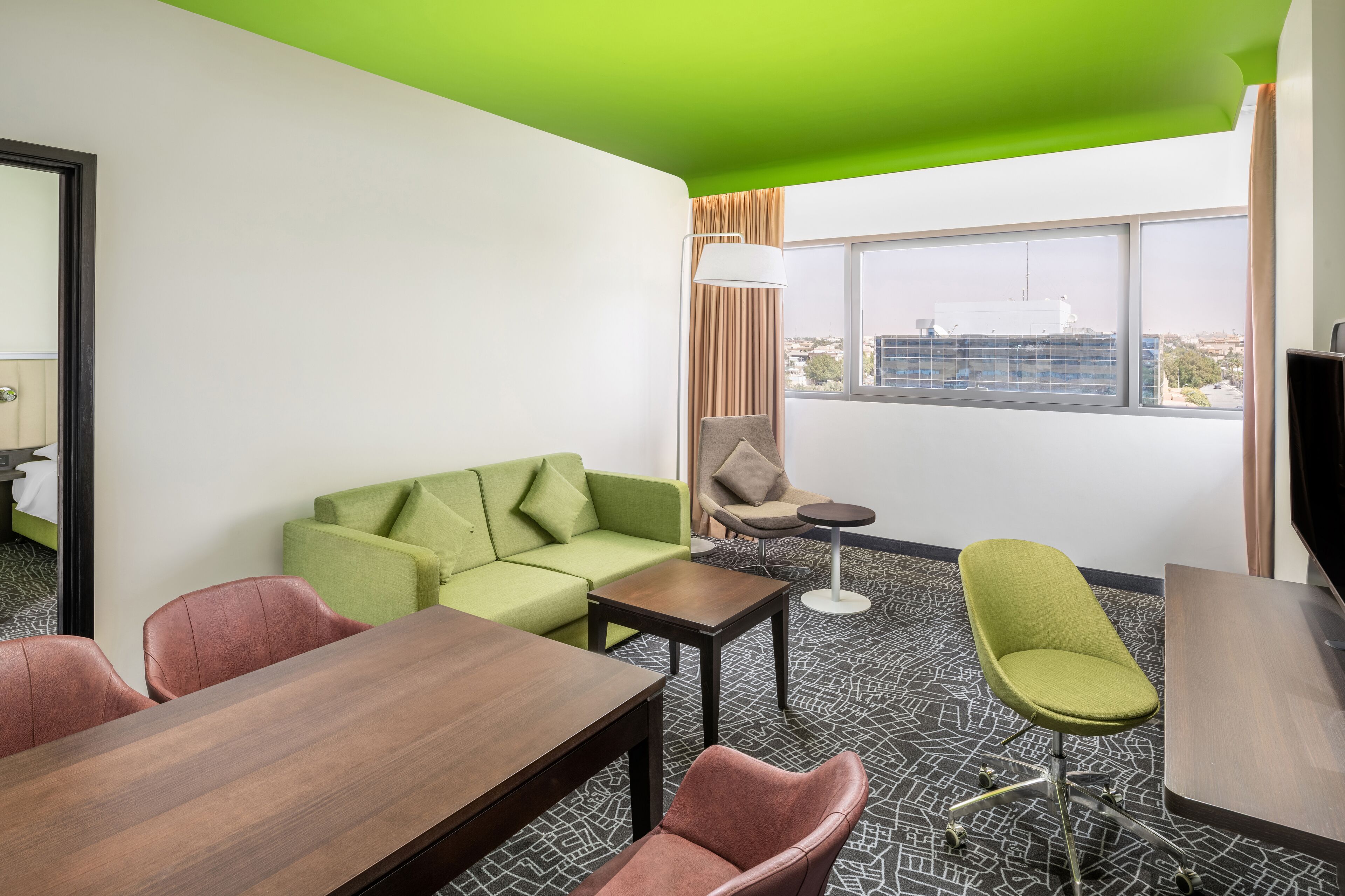 A modern hotel room with a green ceiling, a large window with a city view, a green sofa, a wooden table, and two chairs.