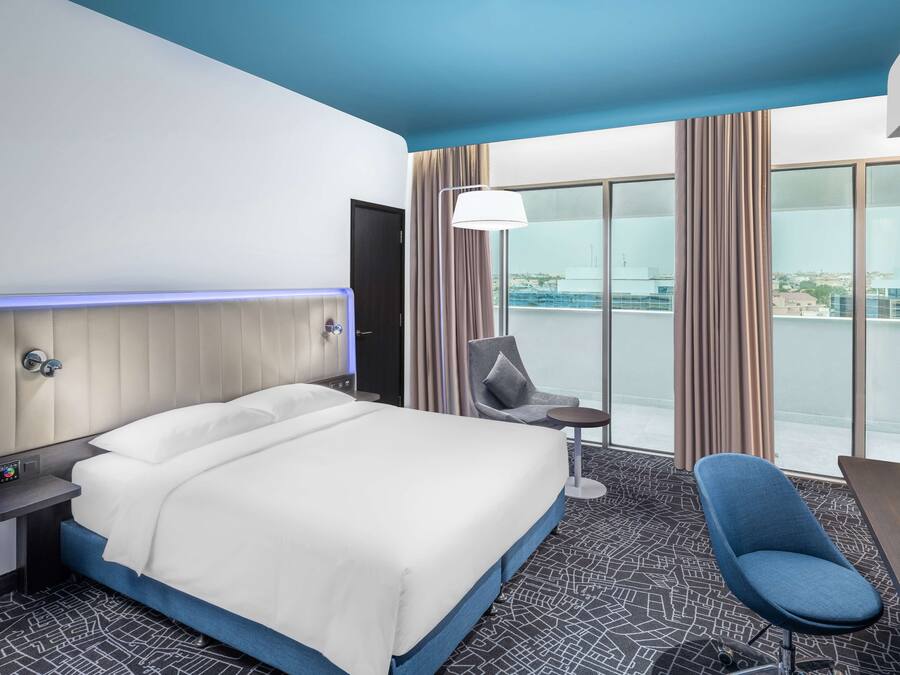 A modern hotel room with a large bed, a desk, a chair, and a view of the city through large windows.