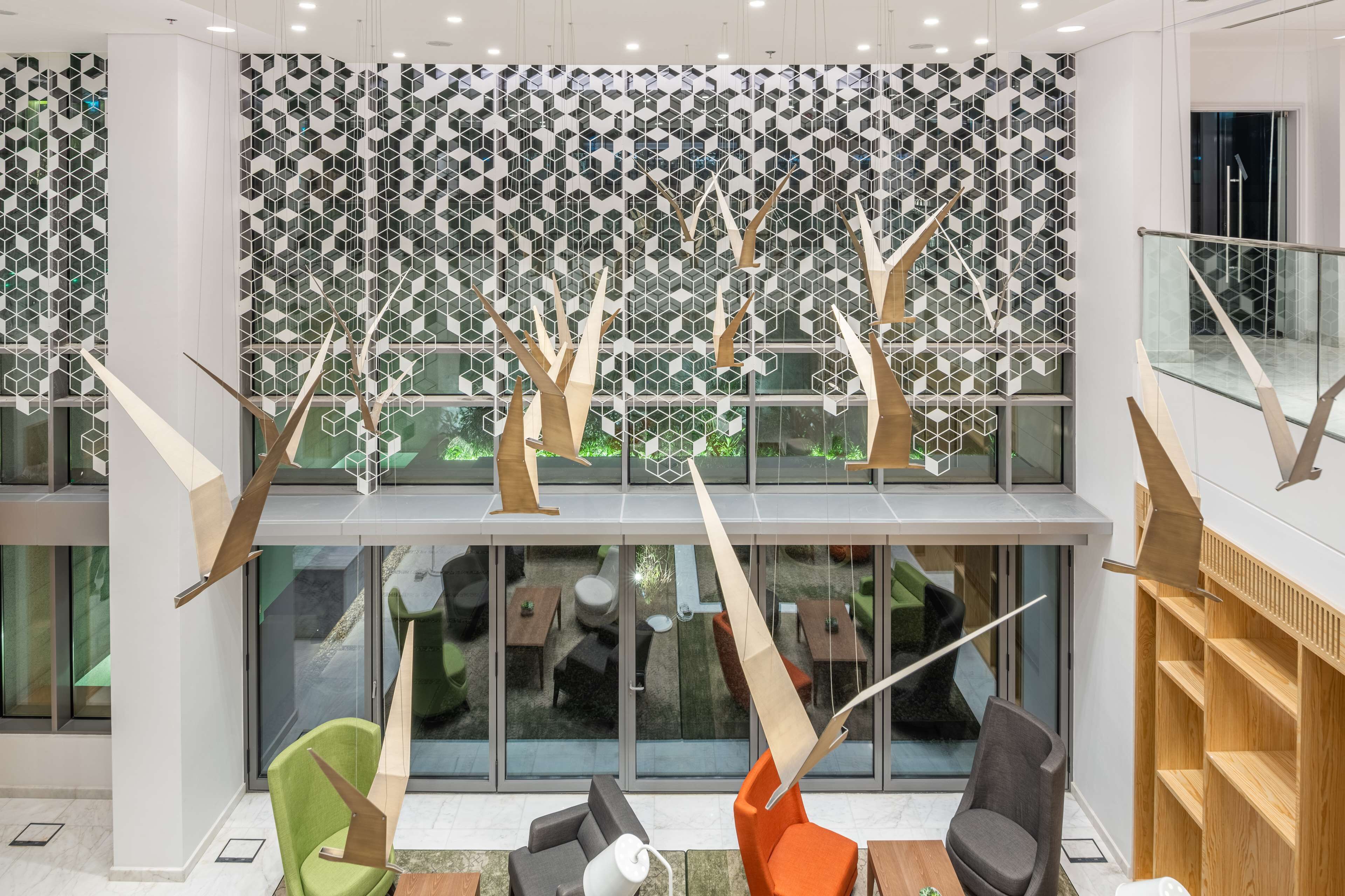 Un interior moderno con un diseño geométrico en la pared, una zona de estar con sillas y mesas, y una escalera con barandillas de madera.
