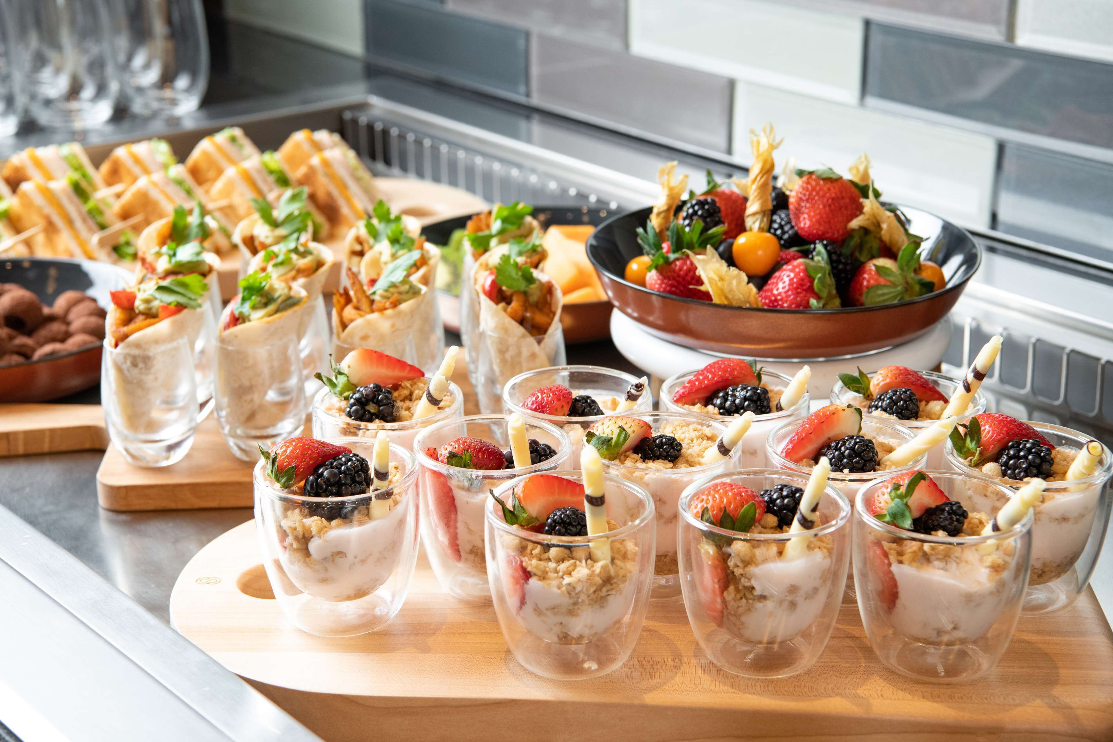 A buffet with various food items including sandwiches, a bowl of mixed fruits, and dessert cups with strawberries and blackberries.