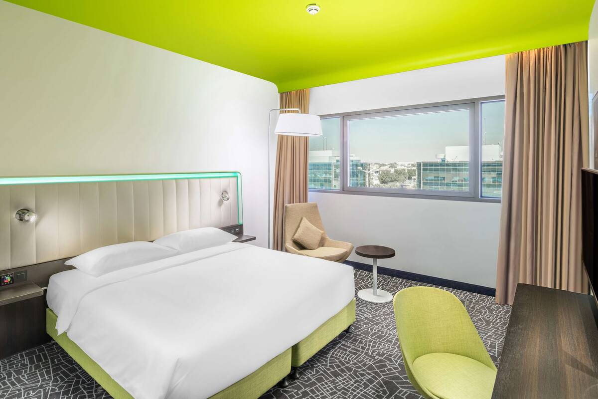 A hotel room with a large bed, a chair, a small table, and a window with curtains.