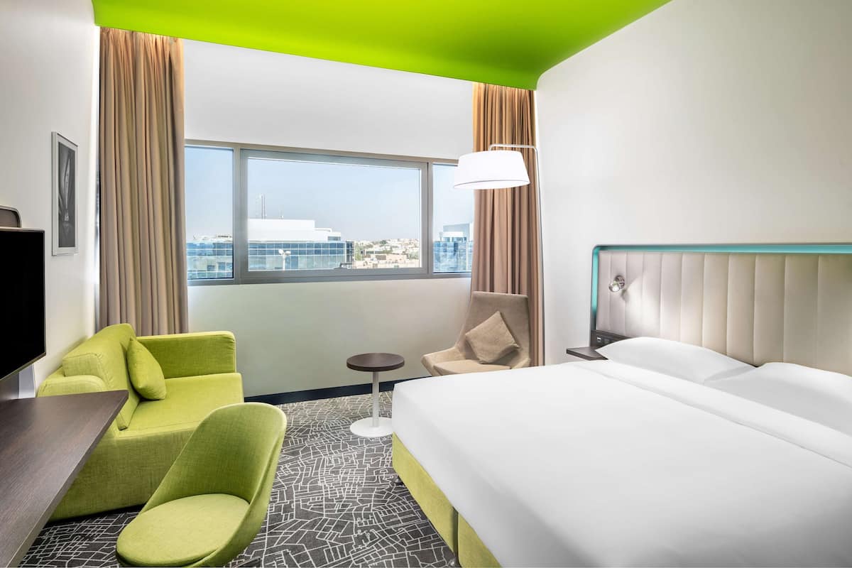 A hotel room with a bed, a green armchair, a small round table, and a view of the city through large windows.