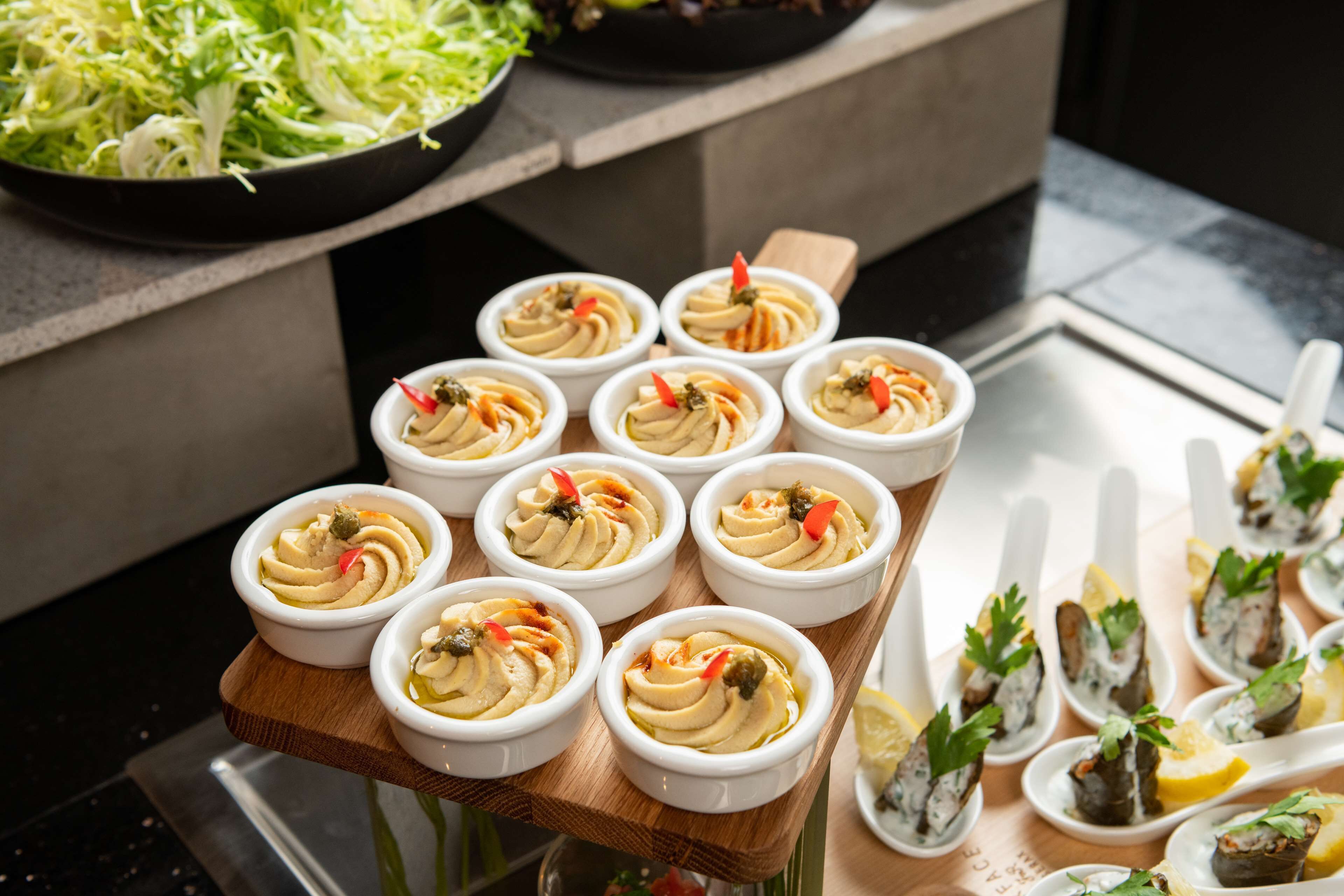 A tray of small white bowls with a creamy dish, garnished with red chili and herbs, on a wooden board.