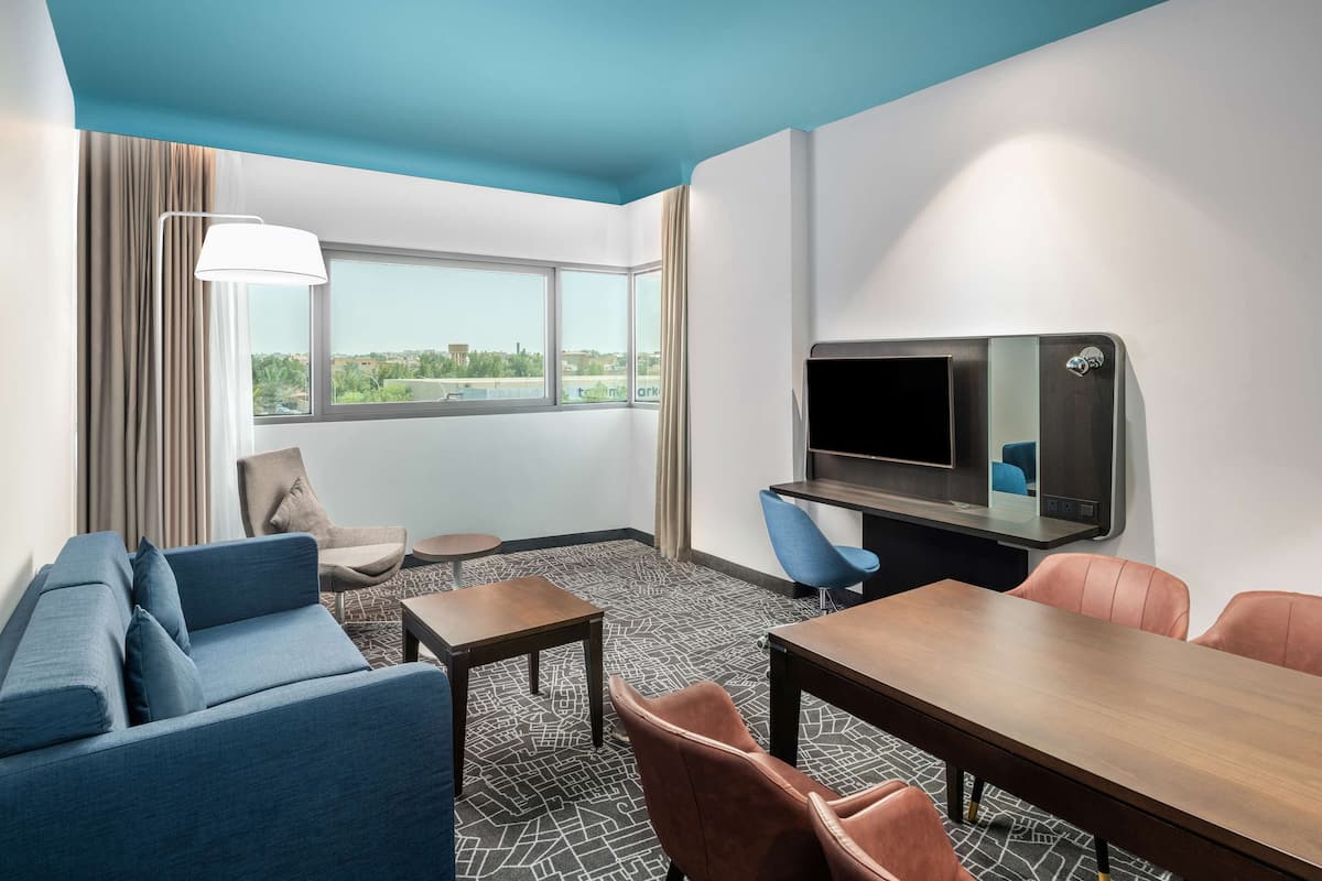 A hotel room with a blue ceiling, a large window, a sofa, a table, and a TV.