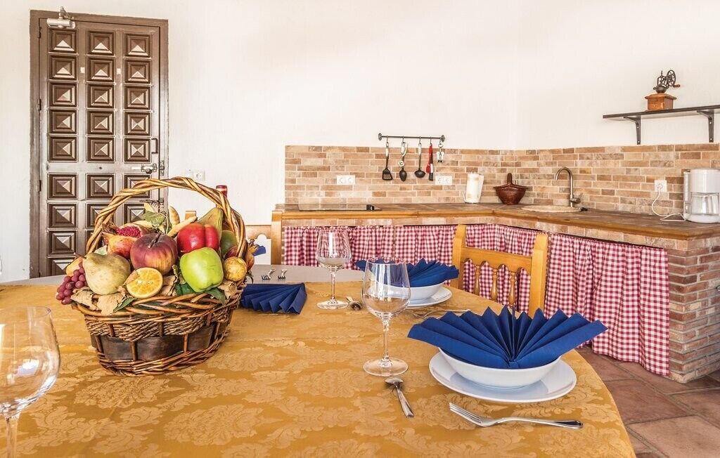 Un coin repas avec une table dressée pour deux personnes, un panier de fruits et une cuisine avec un mur arrière en briques et des ustensiles de cuisine suspendus.