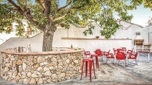 Zona de bar al aire libre con mostrador de piedra, taburetes rojos y un árbol que da sombra.