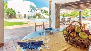 Mesa de comedor con platos, servilletas y una cesta de fruta, con vistas a una piscina y edificios.