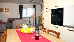 Uma área de refeições com mesa de madeira, cadeiras, toalha de mesa vermelha, uma garrafa de vinho e uma cesta de frutas.