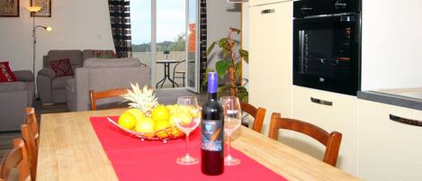 A dining area with a wooden table, chairs, a red tablecloth, a bottle of wine, and a fruit basket.