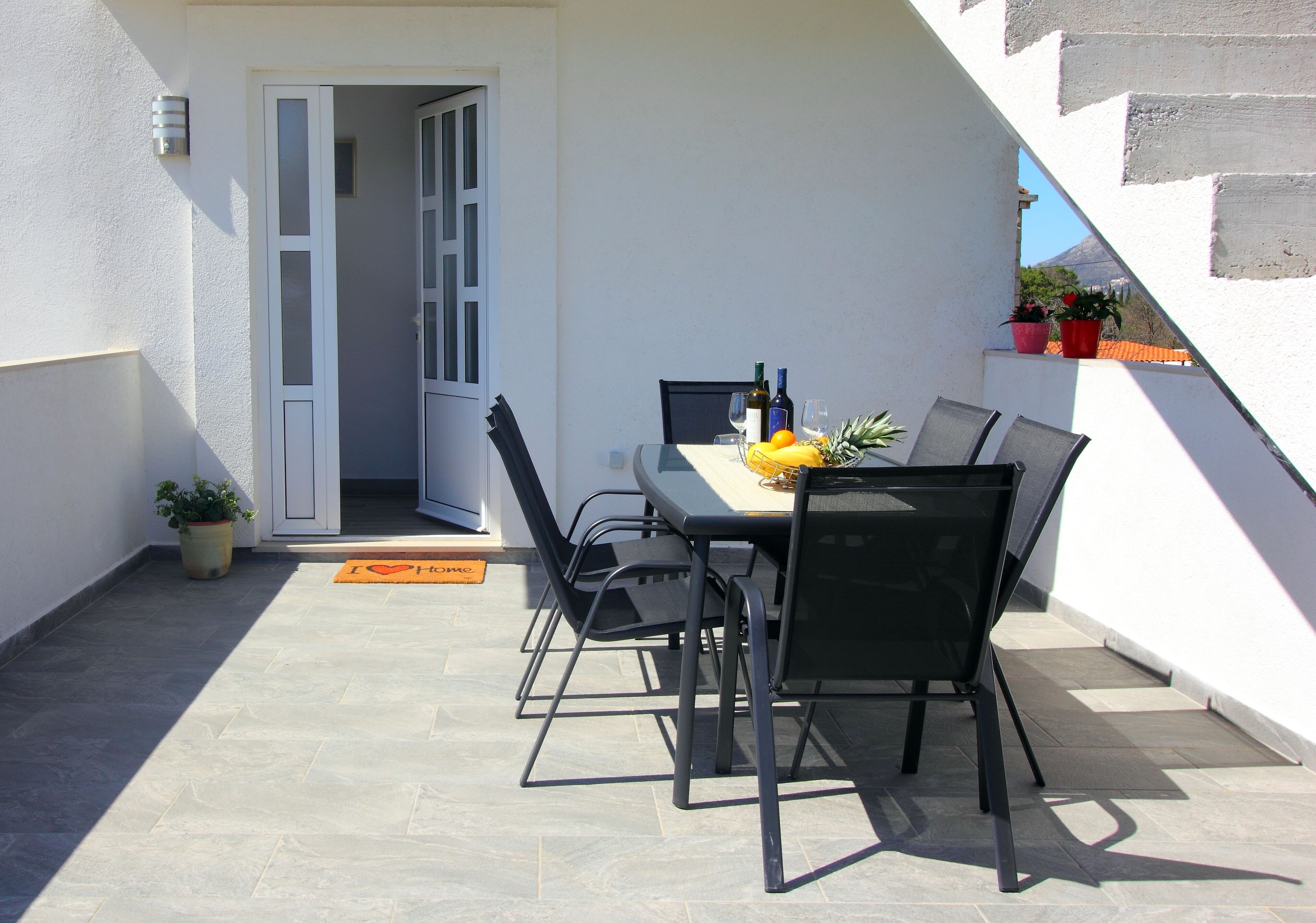 A patio with a table set for two, chairs, a doormat, and potted plants.