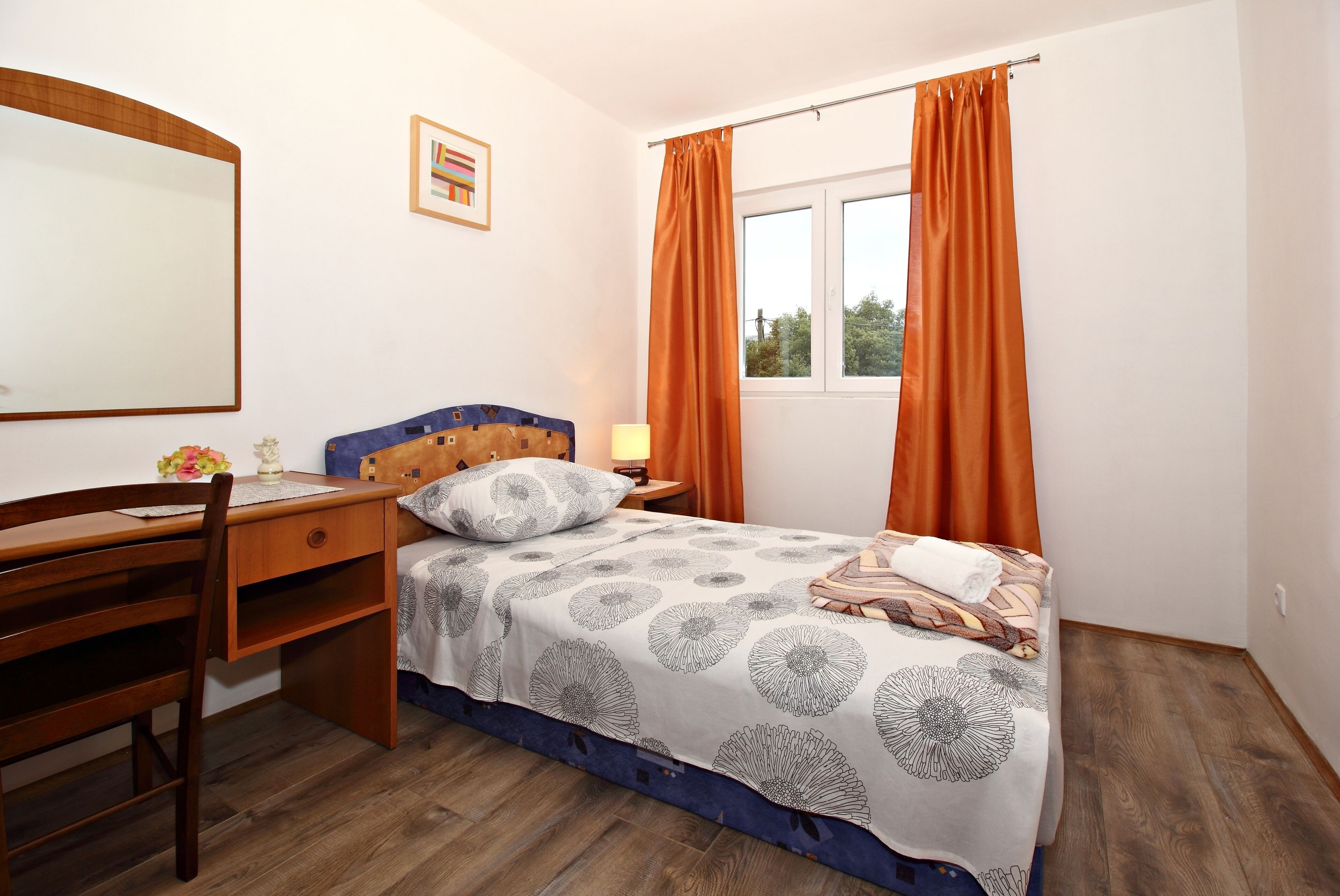 A bedroom with a wooden desk, a bed with a patterned bedspread, an orange curtain, and a window with a view of trees.