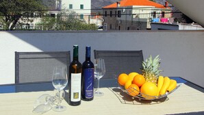 Uma mesa com duas garrafas de vinho, dois copos de vinho e uma cesta de frutas com laranjas e uma abacaxi numa varanda com vista para uma área montanhosa.