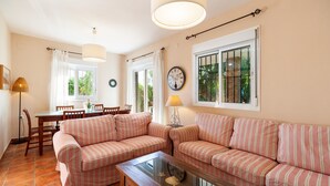 A living room with striped sofas, a glass coffee table, a dining area with chairs, a lamp, and a clock on the wall. - Holiday Home "La Buganvilla" For Families With Garden and Large Pool (Conil de la Frontera)