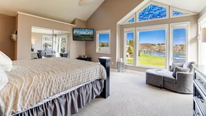 Una camera da letto con un letto grande, un divano grigio e vista su un lago attraverso ampie finestre.