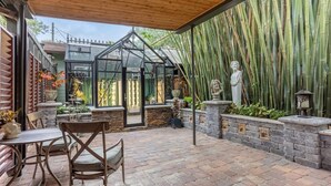 Eine Terrasse mit Pflasterboden, einem Holztisch und Stühlen, einem Glasgewächshaus und Bambuspflanzen.