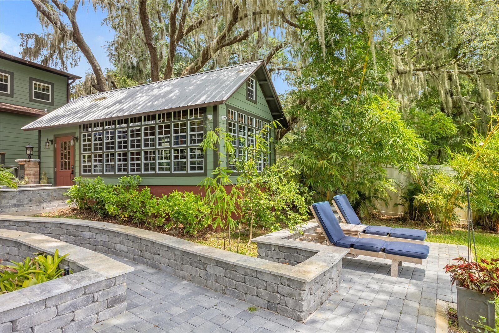 A green house with a large window, a stone patio, and two blue lounge chairs.