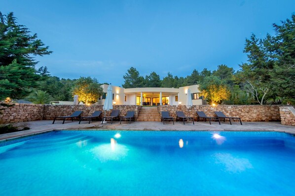 A pool with lounge chairs, a stone wall, and a building with lights.