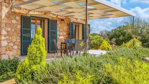 Een stenen huis met een overdekte patio, zitplaatsen buiten en weelderige begroeiing.