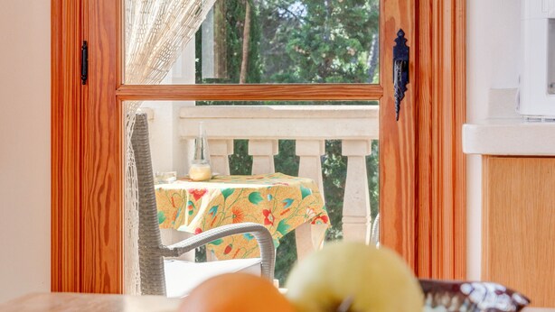 Uma área de refeições com uma mesa posta para dois, uma janela com vista para vegetação e uma cesta de frutas sobre a mesa.