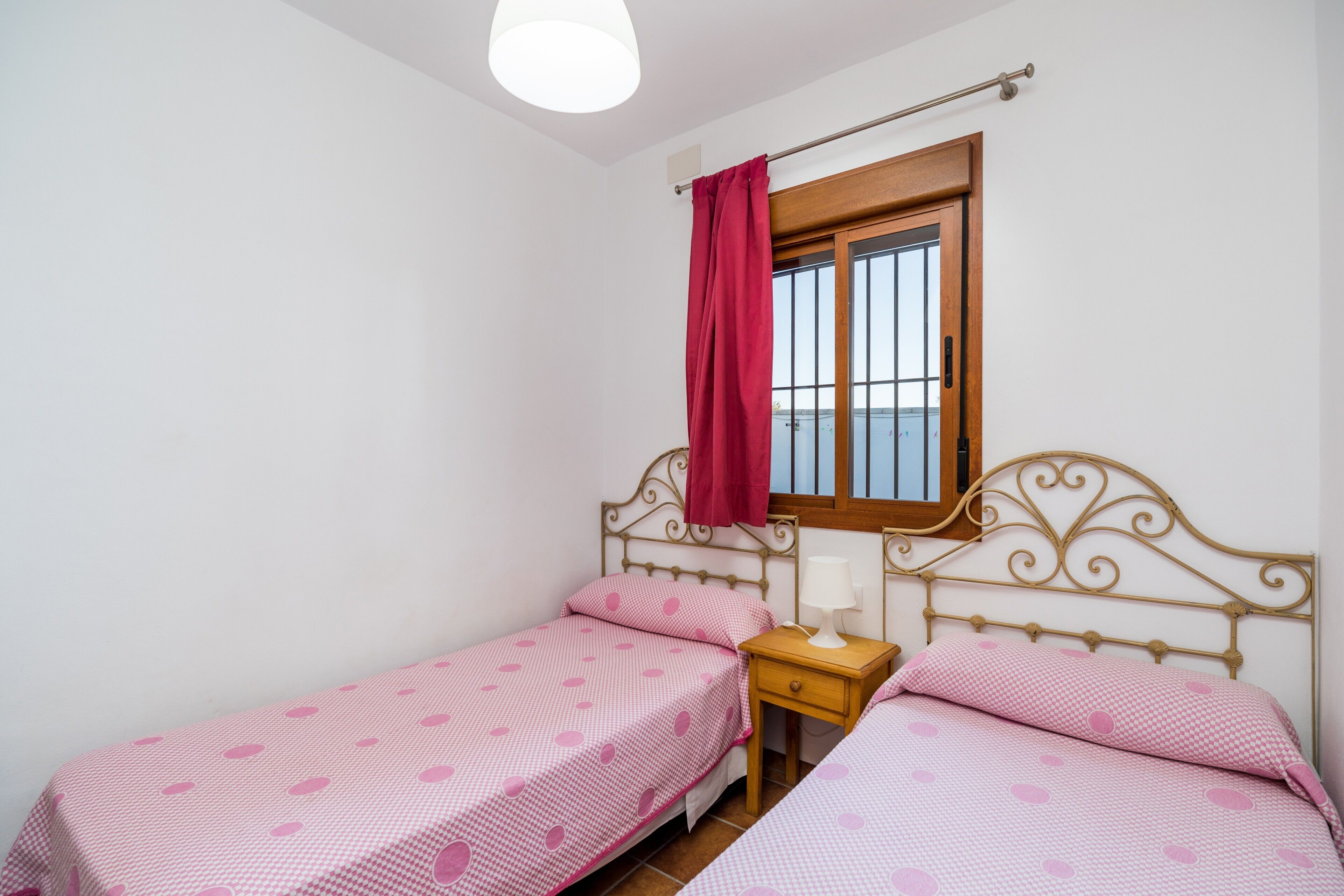 A small bedroom with two single beds, a nightstand, a window with a curtain, and a ceiling light.