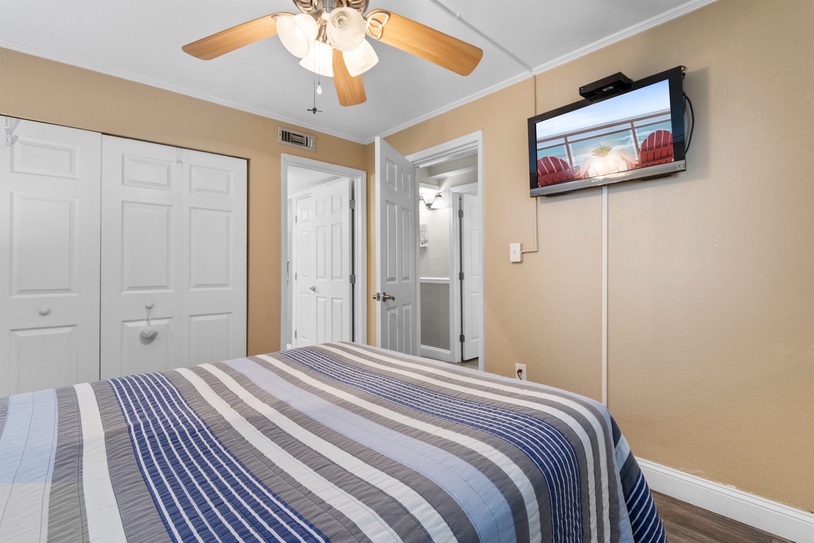 Een slaapkamer met een bed, een plafondventilator, een aan de muur bevestigde televisie en een deur naar een andere kamer.