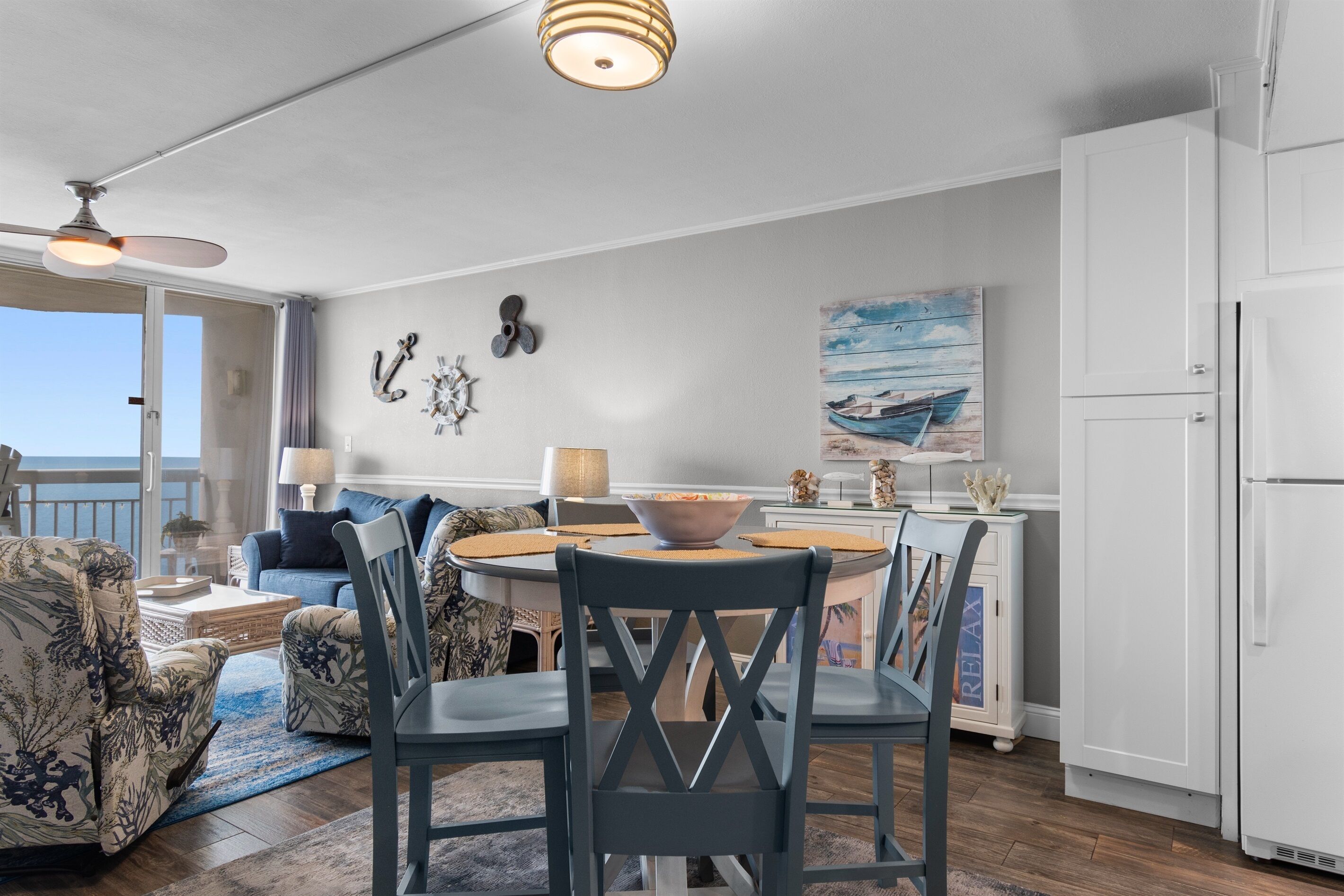 Een eetgedeelte met een ronde tafel, stoelen en een fruitschaal. Er zijn decoratieve wandversieringen en een schilderij van een boot.